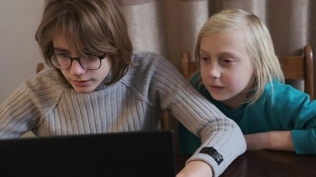 Teenage boy and a young brother are sitting at a table, captivated by something entertaining on a laptop, their expressions shifting in slow motion