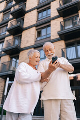 Vertical portrait of cheerful beautiful senior couple with gray-haired in white shirts standing on city street, happily browsing using smartphone, radiating love and joy. Concept of active aging.