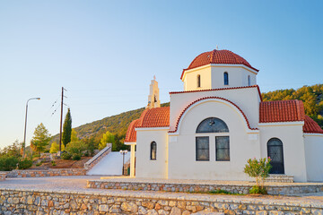 A small, traditional Greek Orthodox church with a red-tiled dome and white walls, set against a backdrop of green hills and a clear blue sky.