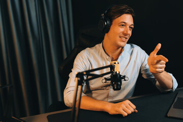 Host channel smart broadcaster talking on live social media application searching on laptop, broadcasting streamer wearing headphone using mic at modern home office studio in warm lighting. Pecuniary.