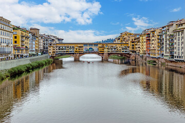 Medieval Florence showcases its stunning architecture with colorful buildings lining the Arno River, where boats glide smoothly along the reflective waters under a bright sky.