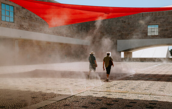 Two people are enjoying a refreshing mist as they walk through the courtyard of centro cultural de belem in lisbon, portugal, under a vibrant red shade on a sunny day