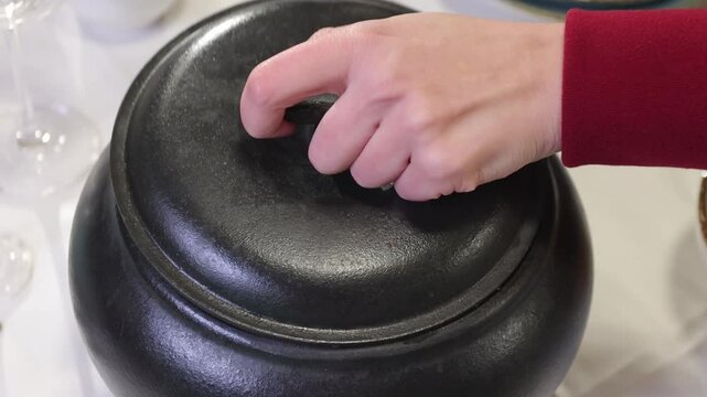 slow-motion of woman's hand opening a black pot, reveals Ossobuco inside it, on a white table