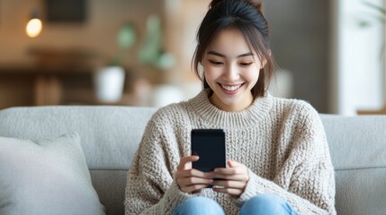  Young Asian woman smiling while using a smartphone at home, sitting on a sofa in the living room with copy space for text 