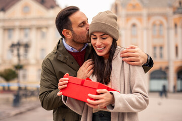 Mid adult Caucasian male delighting his wife with a present, both dressed warmly, outdoors in a historic city setting.