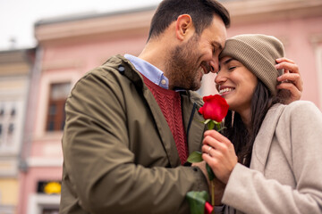 Caucasian couple expresses joy while walking on a city street. Man and woman wear cozy winter attire, holding a red rose.