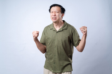 Fototapeta premium An Asian man in a green shirt displays an intense and frustrated expression with clenched fists and scrunched face, conveying anger or determination, against a plain white background
