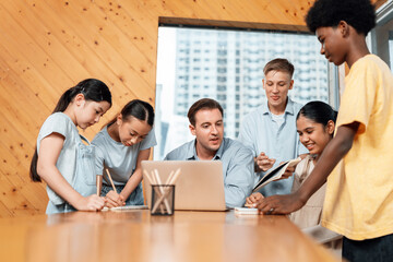 Teacher sitting and giving a lecture on lesson to children in classroom using laptop, talking to...