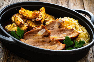 Tasty roast ribs with baked potatoes and sauerkraut salad in frying pan on wooden table