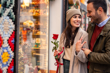 A mid adult Caucasian couple delight in window shopping for Valentine's Day. The woman holds a red rose, wearing a cozy hat and coat, while the man embraces her.