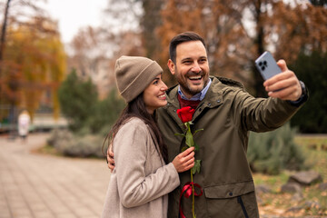 Caucasian mid adult couple, smiling and happy, taking a selfie with a smartphone in a public park. The man holds a red rose, both dressed in casual winter attire.