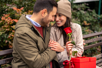 Fototapeta premium Mid adult Caucasian man surprises cheerful woman with a red rose and gift in a public park. Both dressed warmly; man in a green jacket, woman in a beige coat and hat.