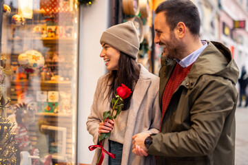 A mid adult Caucasian couple happily explores a charming storefront, the woman holding a single red rose.