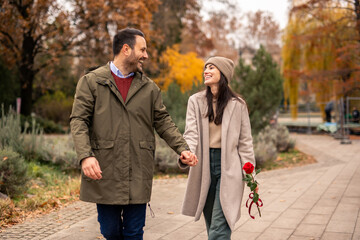 Fototapeta premium Caucasian couple, both dressed in cozy winter attire, leisurely walk hand in hand in a public park. The woman holds a red rose, symbolizing Valentine's Day.