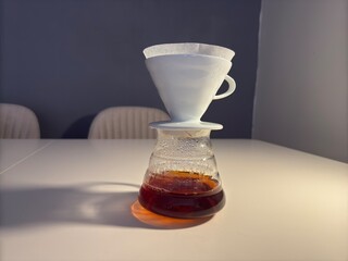A minimalist pour-over coffee setup featuring a ceramic dripper, paper filter, glass carafe, and a black gooseneck kettle on a white table.