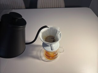 A minimalist pour-over coffee setup featuring a ceramic dripper, paper filter, glass carafe, and a black gooseneck kettle on a white table.