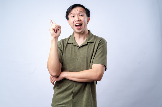 An Asian man in a casual green shirt excitedly points upward with one finger, mouth open in expression, standing against a clean white background. The image conveys an idea, realization