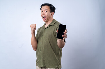 An Asian man in a casual green shirt holds a smartphone forward with an exaggerated surprised expression, standing against a clean white background. The image portrays surprise, presentation