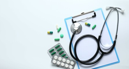 Stethoscope with pills and clipboard on light background, top view