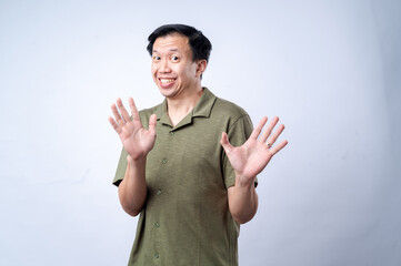 An Asian man in a green polo shirt reacts with an exaggerated surprised expression, raising both hands near his face, conveying excitement or shock, against a plain white background