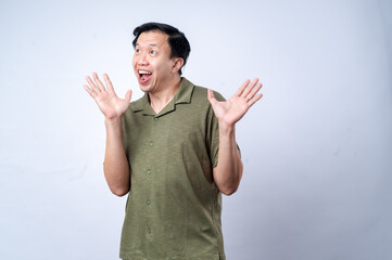 Fototapeta premium An Asian man in a green polo shirt reacts with an exaggerated surprised expression, raising both hands near his face, conveying excitement or shock, against a plain white background