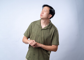 Fototapeta premium An Asian man in a casual green shirt looks to the side with a thoughtful expression, clasping his hands together, standing against a clean white background. The image portrays contemplation