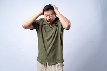 Naklejka premium An Asian man wearing a green shirt, standing against a plain white background, holding his head with both hands and expressing a painful or stressed facial expression, depicting a headache