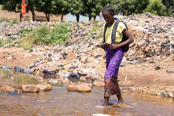 Young African schoolgirl watching her steps as she wades through a small rocky river, symbolizing the lack of infrastructure and public transportation in developing countries