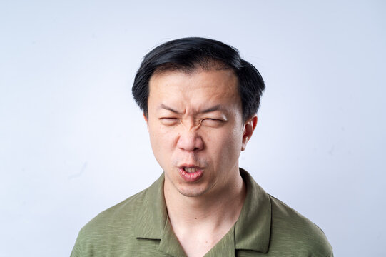 A close-up of an Asian man in a green shirt making a quirky, puckered expression with raised eyebrows and a tilted head, against a plain white background, showcasing a humorous