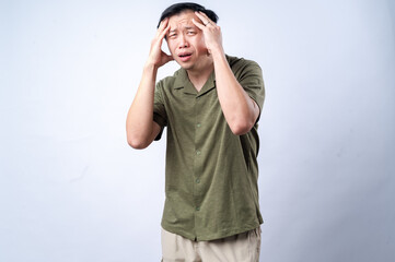 An Asian man wearing a green shirt, standing against a plain white background, holding his head with both hands and expressing a painful or stressed facial expression, depicting a headache