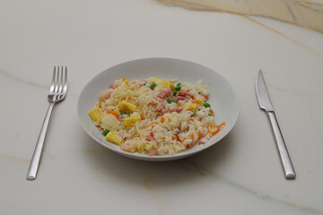 a plate of fried rice on a white marble table, with omelette, peas, ham, carrots and shrimp next to a stainless steel fork and knife
