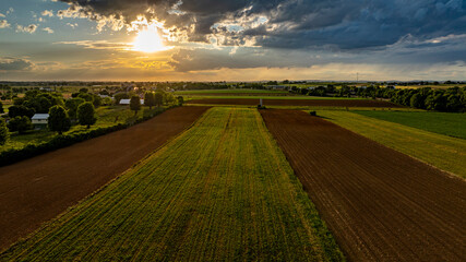 As the sun sets in a vibrant sky, vast agricultural fields stretch across the landscape, dotted with trees and farm structures, creating a serene and picturesque rural scene.