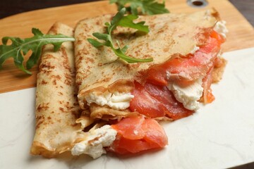 Delicious crepes with salmon, cream cheese and arugula on wooden board, closeup