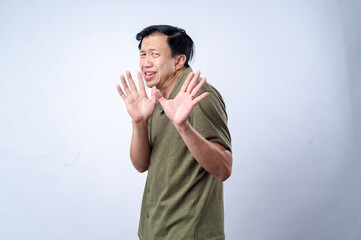 Naklejka premium An Asian man wearing a green polo shirt reacts with a startled expression, raising his hands defensively as if in fear or surprise, against a plain white background, showcasing vivid emotion