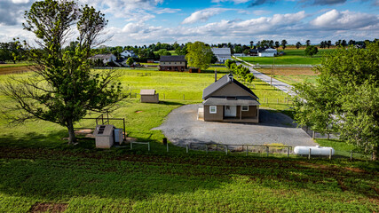 An Amish one room school house sits on a gravel area, surrounded by expansive green fields, barns, and distant structures under a bright blue sky.