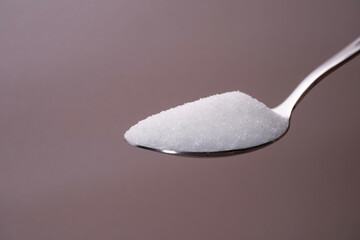 A close up of a single spoon filled with white granulated sugar placed on a neutral brown background.