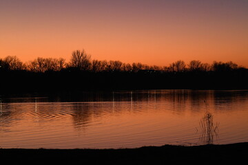 Obraz premium Against the background of a deep lake, an orange sunset flares up in the sky, Light waves on the surface of the water reflect warm colors, Silhouettes of trees on the shore add mystery