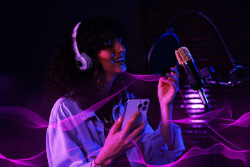 Vocalist with smartphone and headphones singing into microphone in professional record studio....