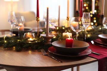 Beautiful dishware with burning candles, fir tree branches and Christmas lights on wooden table indoors, closeup
