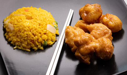 A Chinese dish featuring orange chicken and fried rice, served on a black plate with metal chopsticks.