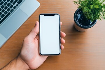 A hand holds a smartphone with a blank white screen beside a laptop on a wooden desk, accompanied by a small potted plant, suggesting modern technology and work.