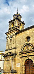 Church facade with a stone bell tower topped by a blue and white dome; detailed carvings on the arched wooden door and window frame; cloudy sky above;