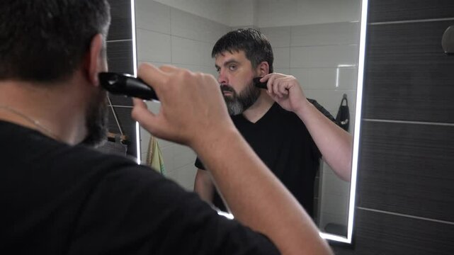 Man shaving his beard in the bathroom. Beard care, electric razor. Reflection of a bearded man in a bathroom mirror.