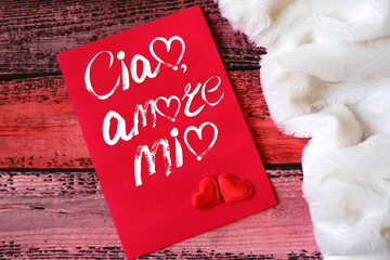 Saying Ciao, amore mio. Text on red card, wooden table. Handwritten message and Translation from Italian Hello my love. Pink background and fluffy warm blanket or plaid © ElenaEmiliya