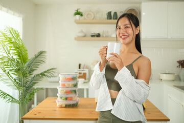 Smiling young sportswoman holding a cup of coffee, standing in her bright kitchen next to a stack of neatly organized meal prep containers. Healthy lifestyle concept.