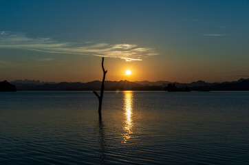 landscape sunset or sunrise on lake sea with golden blue sky or orange sun light on twilight and silhouette mountain range with dry tree trunk or log in water at pom pee vajiralongkorn dam in thailand