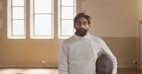 Fencer in white uniform holding helmet, standing confidently in bright room