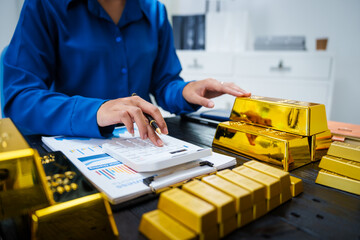 An Asian businesswoman sits at her desk analyzing gold bar prices, buying and selling gold, trading gold stocks, monitoring charts, and studying currency trends in an uptrending stock market.