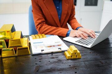 An Asian businesswoman sits at her desk analyzing gold bar prices, buying and selling gold, trading gold stocks, monitoring charts, and studying currency trends in an uptrending stock market.