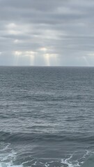 Obraz premium Sunlight beams cut through a cloudy sky, illuminating the calm ocean surface below.
